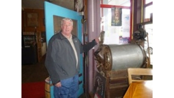 Man with gray jacket and blue jeans next to gray machine inside a building with a blue door.