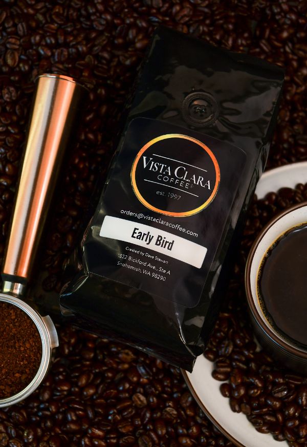 Bag of Vista Clara Early Bird coffee surrounded by beans, with a copper grinder and fresh coffee in a cup.