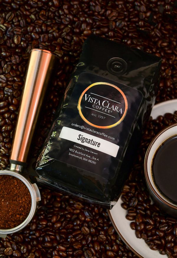 Bag of Vista Clara  Signature coffee surrounded by beans, with a copper grinder and fresh coffee in a cup.