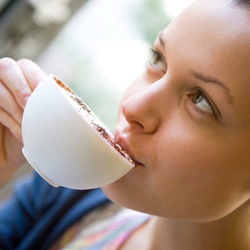 A young woman with a relaxed smile, holding a ceramic coffee cup and taking a sip.